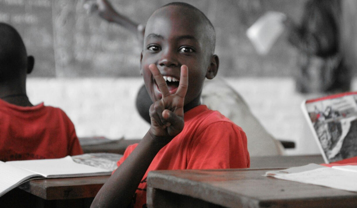 Écolier africain souriant — espoir et paix grâce à l’éducation Jeune garçon en uniforme rouge faisant le signe de la paix dans une salle de classe en Afrique — accès à l’éducation solidaire.